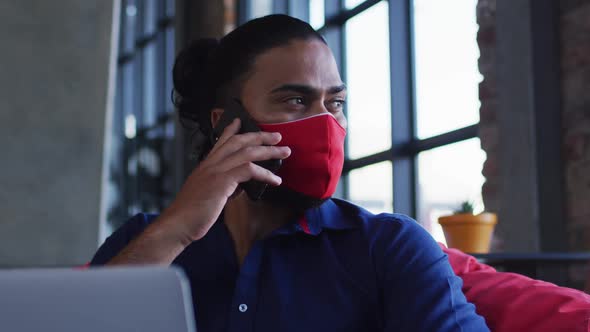 African american man wearing face mask sitting in cafe talking on smartphone using laptop alt