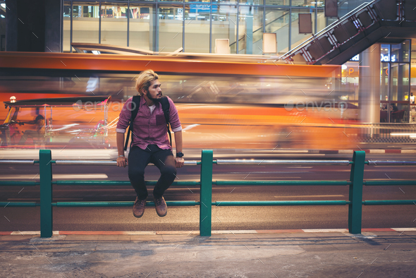 Hipster bearded man sitting on railing with car motion background while  traveling at night. Stock Photo by Johnstocker