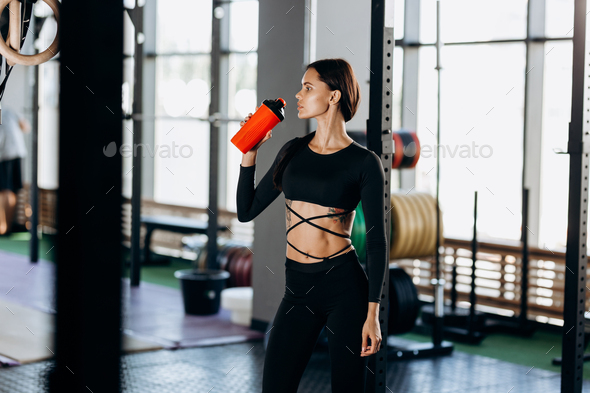Slim dark-haired girl dressed in black sportswear drinks water in the gym near the sport equipment