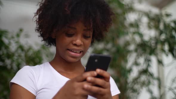 Closeup Lowangle View of Cheerful AfricanAmerican Young Woman Holding Smartphone in Hands Enjoying alt
