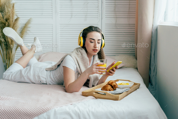 girl is lying on her bed, having breakfast and listening to music on headphones and chatting