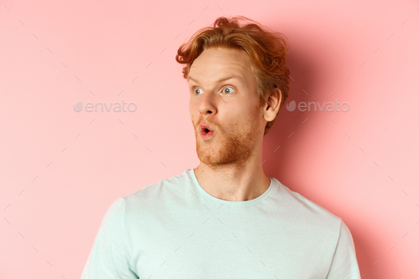Headshot portrait of surprised redhead man with beard, looking left and saying wow, raising eyebrows