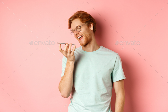 Happy young man with red hair, smiling pleased while record voice message on smartphone, talking to