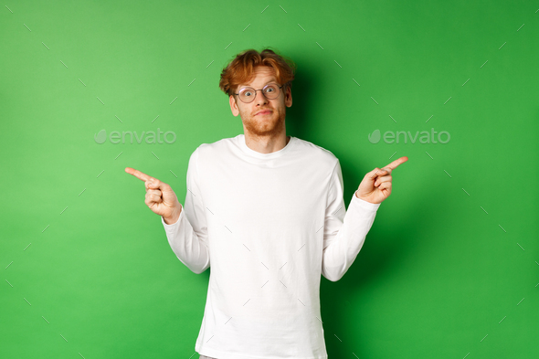Indecisive funny guy with red hair pointing fingers sideways, staring confused at camera and showing