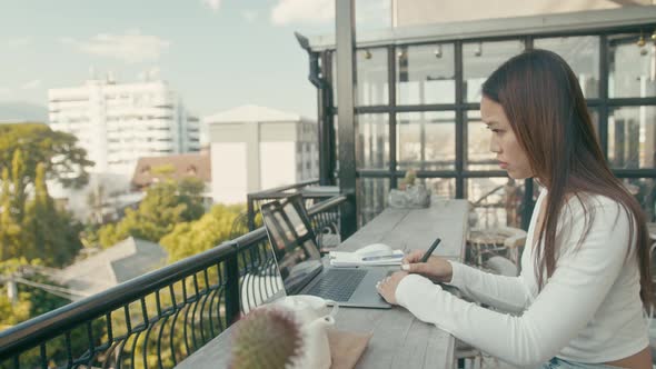 A Young Asian Girl Works at Her Laptop in a Rooftop Cafe in the City alt