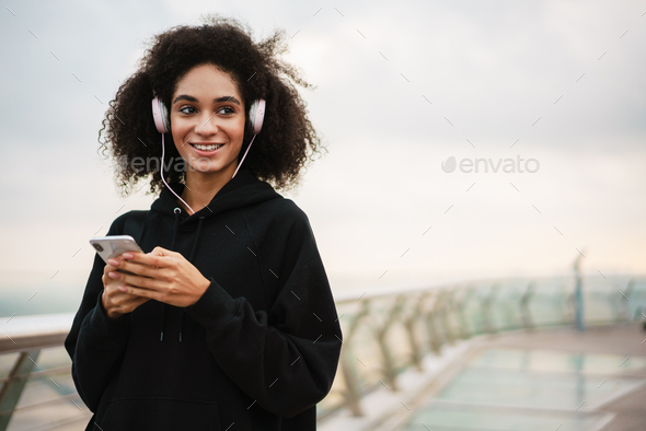 Cheerful athletic sportswoman in headphones using mobile phone