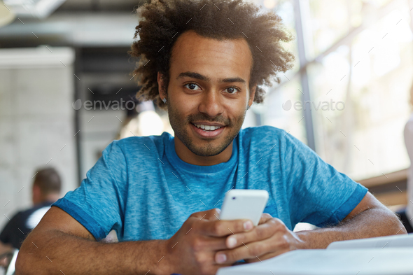 Handsome Afro American guy with head of curly hair sitting in cozy cafeteria holding smart phone dow