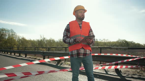 Young Bearded Man Inspector in an Orange Vest and Protective Helmet with Tablet alt