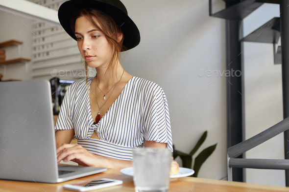 Portrait of elegant lady wearing black hat and blouse sitting in office in front of opened laptop wo