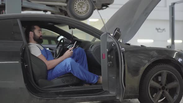 Successful Mechanic Sitting Inside the Car and Verifies Information on His Phone alt