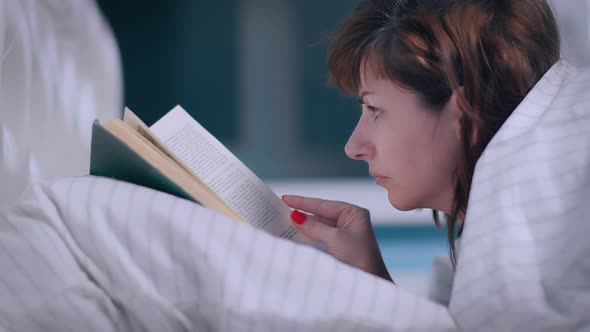 Brunette Woman Covered with a Blanket Lies on the Bed and Reads a Book at Night Cinematic Shot