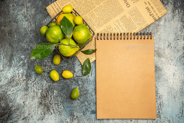 Horizontal view of black basket with fresh green tangerines and kumquats on newspapers and notebook
