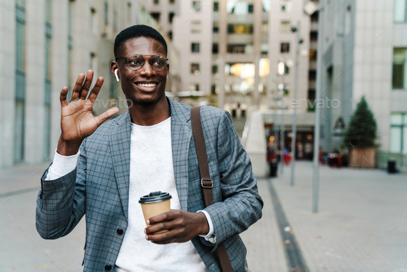 Happy african american man in earphones waving hand while drinking coffee