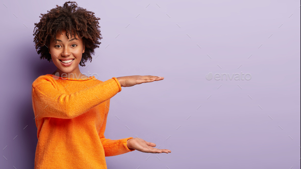 Positive dark skinned girl makes large or big object with both hands, shapes huge box, wears orange