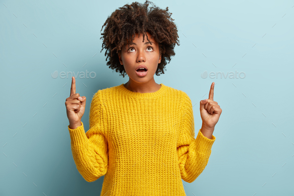 Fascinated dark skinned lady with curly hair, dressed in yellow sweater, points upwards with scared