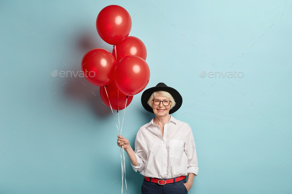 Happy elderly female in festive clothes, holds bunch of air balloons, celebrates anniversary, waits