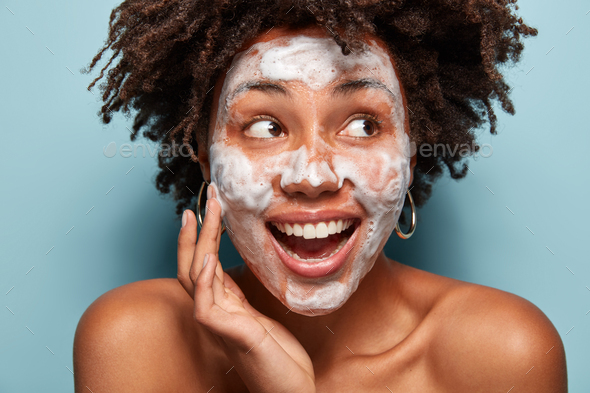 Headshot of happy black woman with foam or foaming cleanser on face, has facial treatments, gazes as