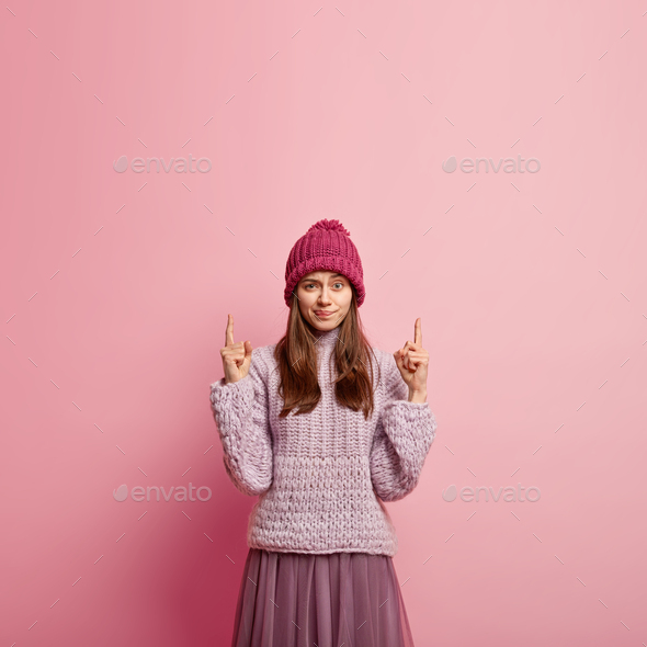 Photo of discontent woman purses lips, has angry expression, dressed in warm clothes, points upwards