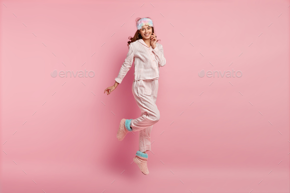 Good morning! Positive young European woman in nightwear, eyemask, jumps in air, has glad expression