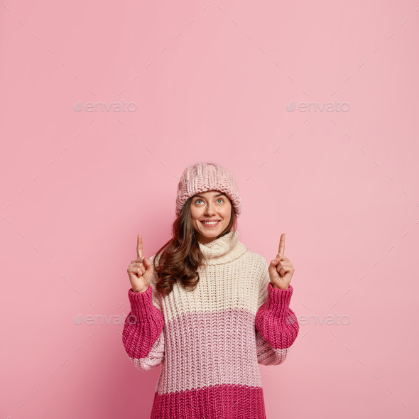Shot of satisfied European woman points upwards with index fingers, wears headgear and oversized jum