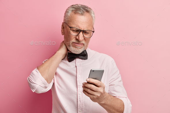 Dissatisfied attractive mature businessman focused in screen of cellular, touches neck, checks email