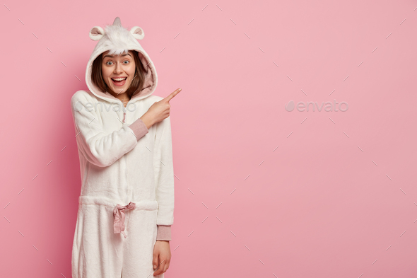 Photo of pleased lovely young woman with satisfied expression, dressed in sleepwear, points aside on