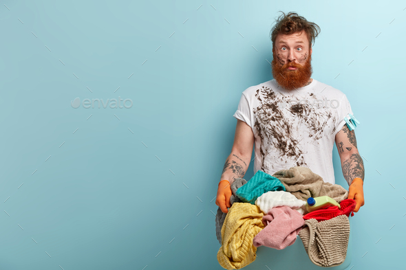 Crazy bearded man crosses eyes, has funny facial expression, dirty clothes and face, carries basin w