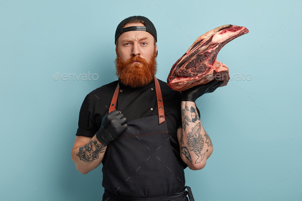 Strict ginger serious male butcher holds very big piece of meat, frowns ...