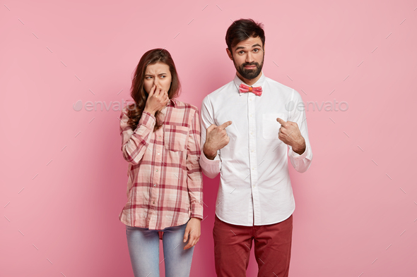 Dissatisfied young woman covers nose as smells stink, unpleasant smell, curious bearded man points a