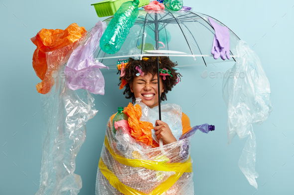 Emotional dark skinned woman stands under plastic umbrella, smirks face and shows white teeth, demon