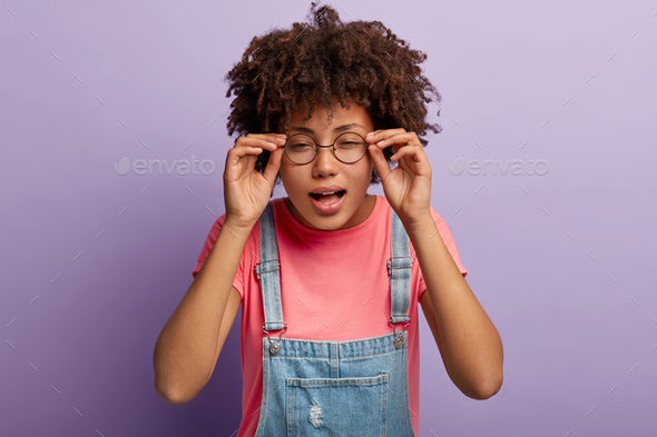 Young Afro girl keeps hands on frame of glasses, tries to concentrate, has bad vision, looks into di