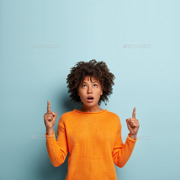 Indoor shot of wondered surprised beautiful black woman points upwards, shows direction above, wears