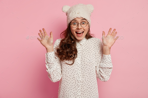 Photo of excited European woman shows palms, feels amazed, wears white hat with ears, polka dot blou