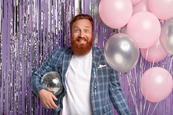 Joyful handsome man throws party after promotion at work, holds disco ball and bunch of air balloons