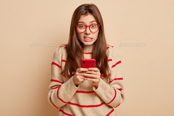 Photo of frustrated displeased woman frowns face, smirks face, holds mobile phone, dressed in sweate