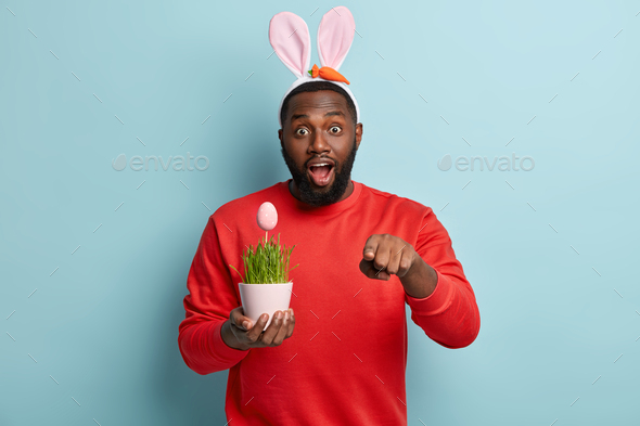 Horizontal shot of stupefied dark skinned plump man points at camera at you, being Easter bunny on s