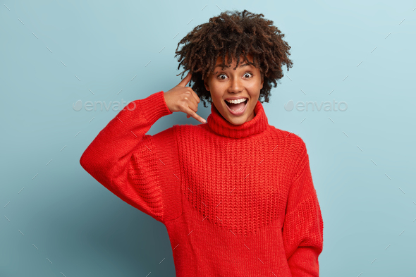 Happy overjoyed Afro American woman shows call me gesture, asks keep in touch, wears warm red sweate