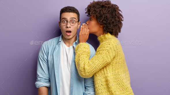 Photo of Afro American woman whispers secret to boyfriend, tells shocking news. Astonished young man