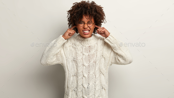 Indoor shot of frustrated woman with dark skin, curly hair, plugs ears, clenches teeth, avoids bad s