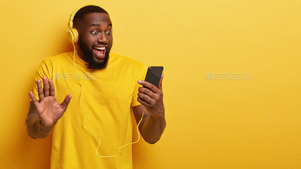 Indoor shot of cheerful guy with dark skin, sings along with favourite song, holds mobile phone, sho