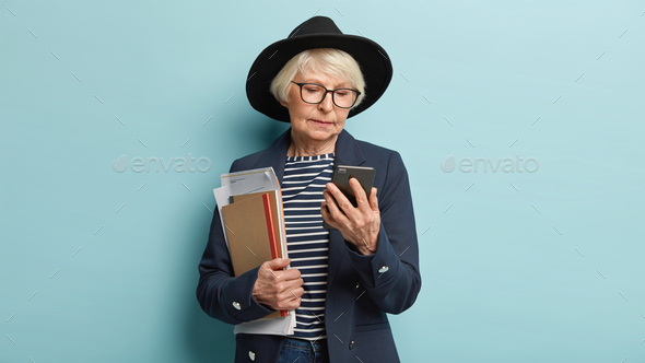 Concentrated elderly headmistress focused in cell phone, holds some business papers, checks email bo