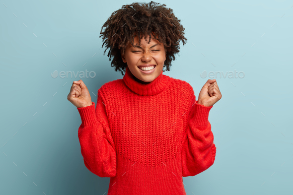 Waist up shot of satisfied black woman raises clenched fists, enjoys her victory, expresses happines