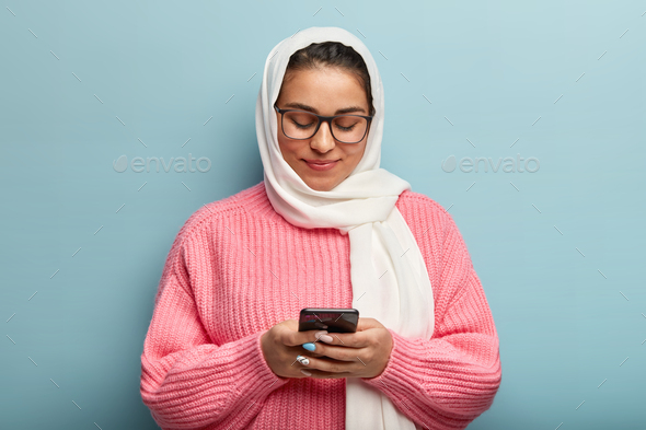 Shot of attractive Arabian woman holds mobile phone, sends text messages and checks email box, focus