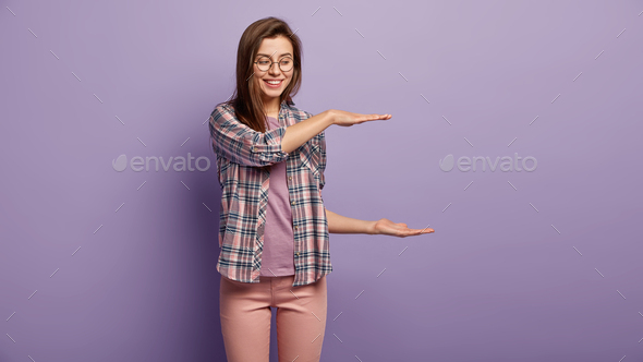 Studio shot of cheerful Caucasian woman measures height of invisible object, demonstrates big item,