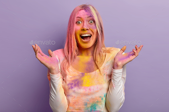 Excited lovely female laughs happily, raises hands, has multicolored face covered with powder paint