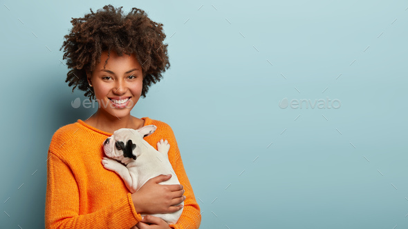 Photo of lovely affectionale female dog host with satisfied expression, carries french bulldog puppy