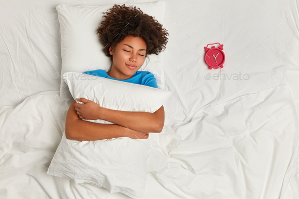 Indoor shot of satisfied dark skinned woman has rest in bed, sleeps well, focuse on red alarm clock,