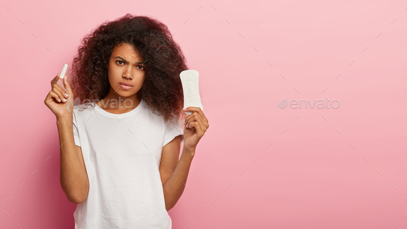 Serious unhappy curly haired lady holds tampon and sanitary pad, thinks which kind of protection bet
