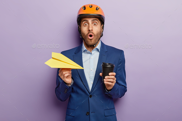 Scared male technician in safety helmet stares with omg expression, holds hand made paper plane and