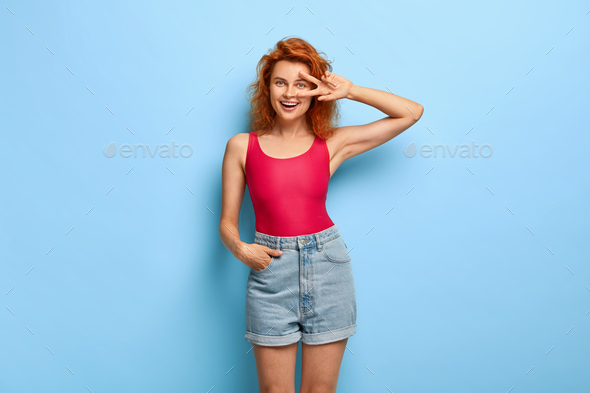 Positive red haired young woman makes peace gesture, feels lucky, has fun, keeps one hand in pocket